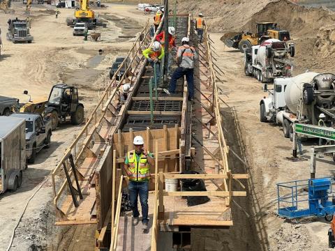 A construction site with multiple workers wearing hard hats and safety vests on a bridge structure. They are working with wooden planks and metal reinforcements. Various construction vehicles, including a cement truck, excavator, and a pickup truck, are visible in the background. The site appears to be in a sandy or dirt area, with machinery and equipment around the perimeter.