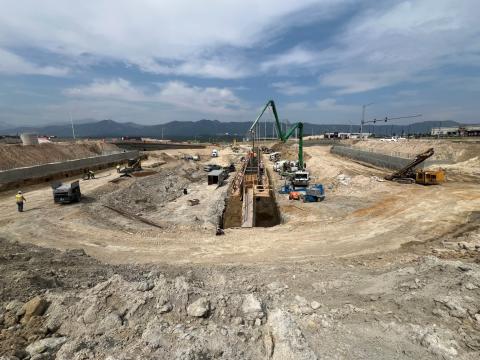 A wide-angle view of a construction site with a deep excavation in progress. The image shows several construction workers in high-visibility vests and helmets working around the site. There are various construction vehicles, including excavators, cement mixers, and trucks, operating within the excavation area. A large concrete pumping truck with an extended arm is visible, along with temporary wooden structures and scaffolding. In the background, there are mountains under a cloudy sky, and several buildings