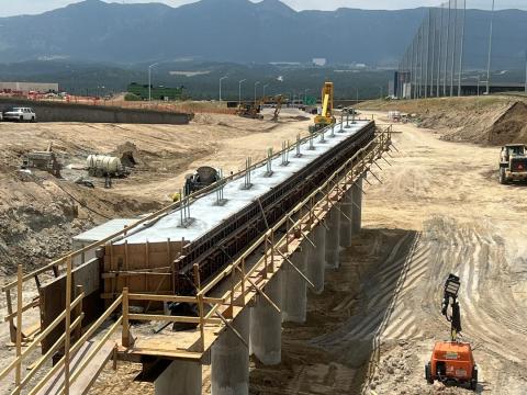 An image of a construction site showing a partially built bridge or elevated structure supported by a series of concrete columns. The structure is lined with temporary wooden scaffolding and handrails. Construction equipment, such as an excavator and a lift, are positioned around the site. Workers in high-visibility gear are seen performing various tasks. The surrounding area is an expansive dirt landscape with additional machinery and construction materials scattered throughout. A distant view of a highway