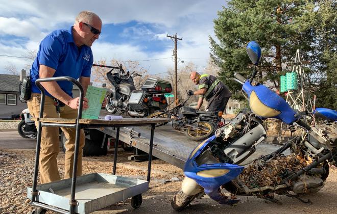 A public works employee looks at a motorcycle covered in leaf litter.
