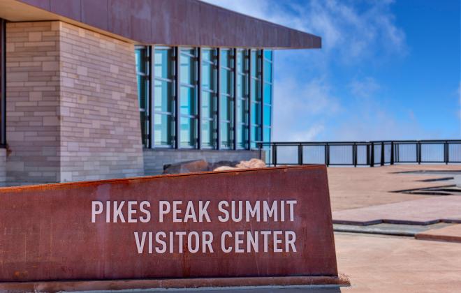 Pike Peak Summit Visitor Center sign. Outdoor viewing platform and wall of windows on the visitor center in the background.