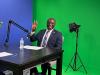 Mayor Yemi smiles at the camera in front of a green screen and holds up four fingers.